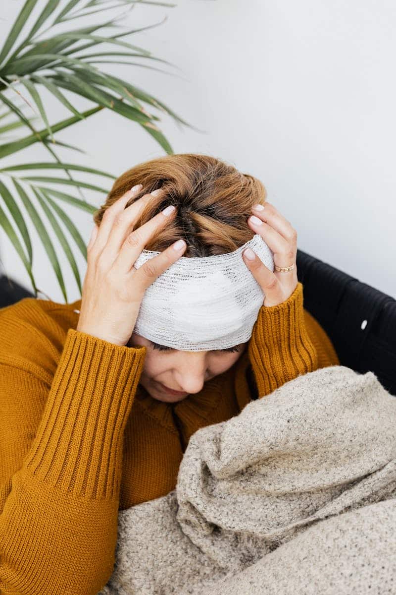 Woman with a bandage holding her head, expressing pain, indoors near a potted plant.