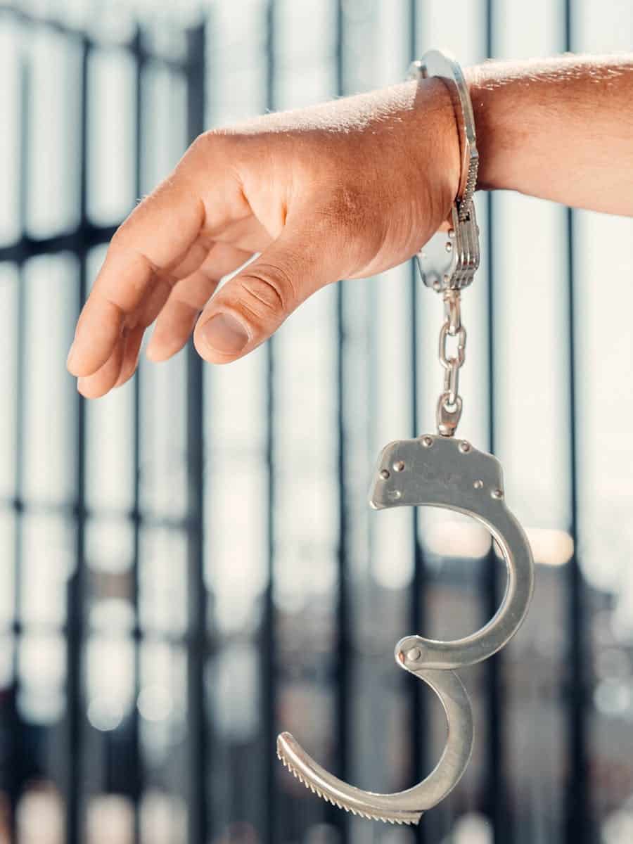 A close-up, conceptual shot of a handcuffed hand with one open handcuff against blurred bars.