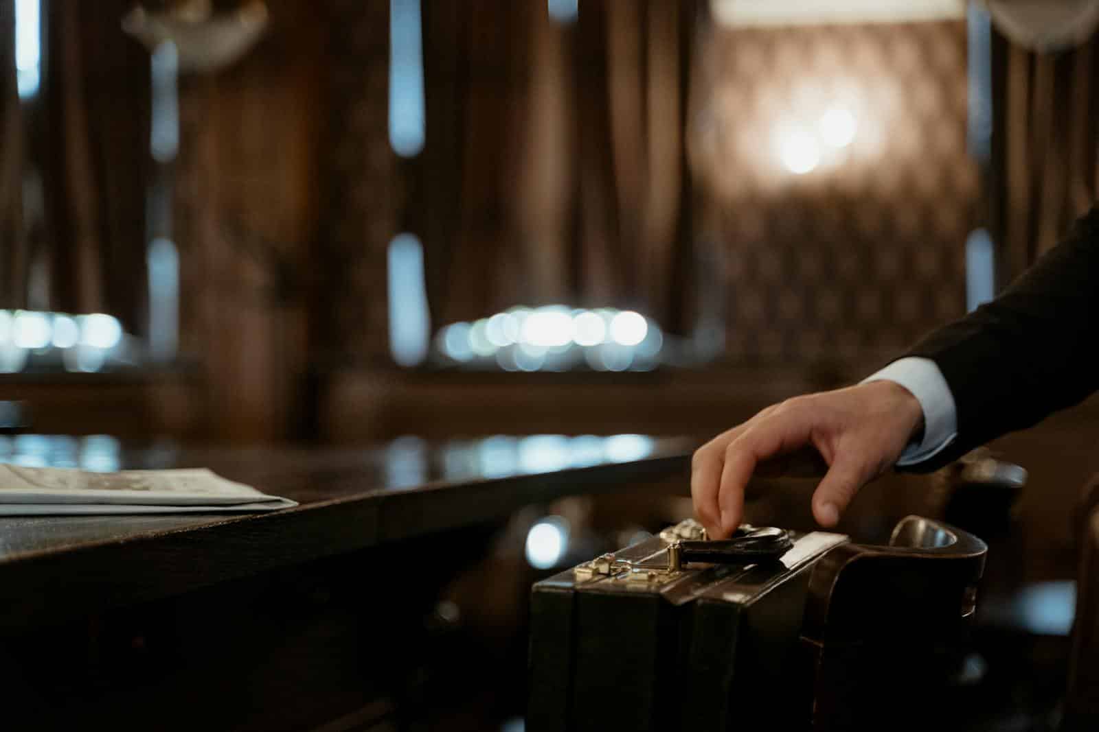 A close-up shot captures a hand holding a black briefcase in a dimly lit elegant room with a blurred background.